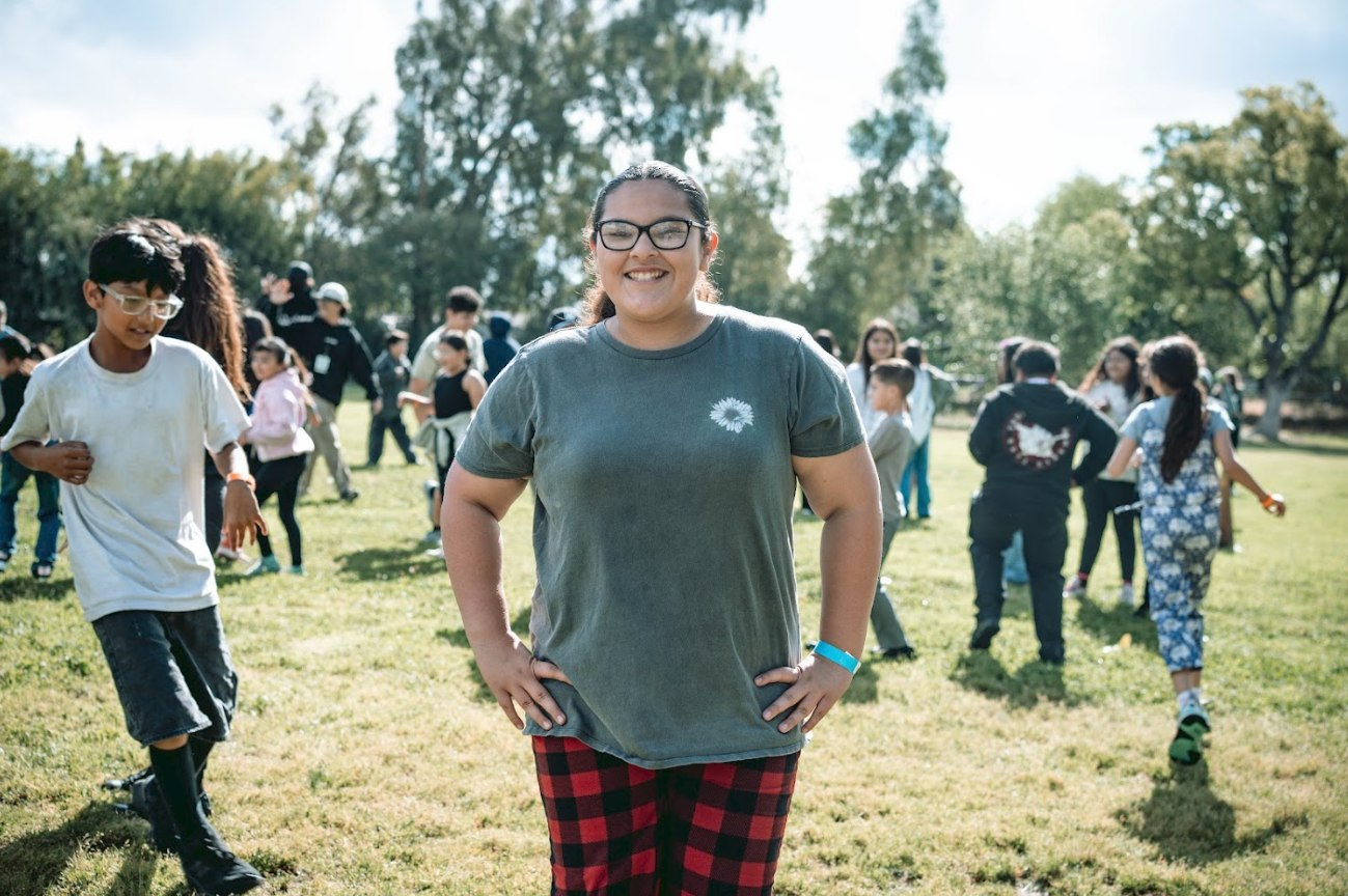 A woman standing on grass with hands on hips, smiling, in a park during daytime, surrounded by children and adults, with trees in the background.