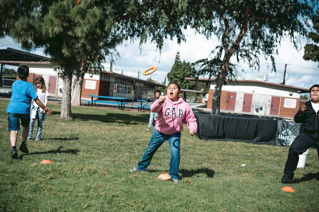 Kids playing disc golf outdoors, with one girl in a pink hoodie throwing a disc on a grassy field under trees, other children watching and playing nearby, school buildings in the background.
