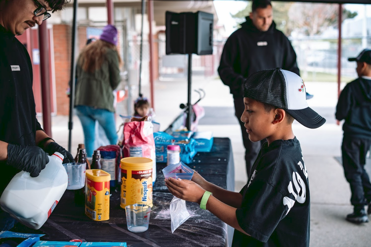 A young boy with a black and white cap and black T-shirt receives ingredients for a science experiment at a table with baking supplies and liquids, overseen by adults, in an outdoor covered area.