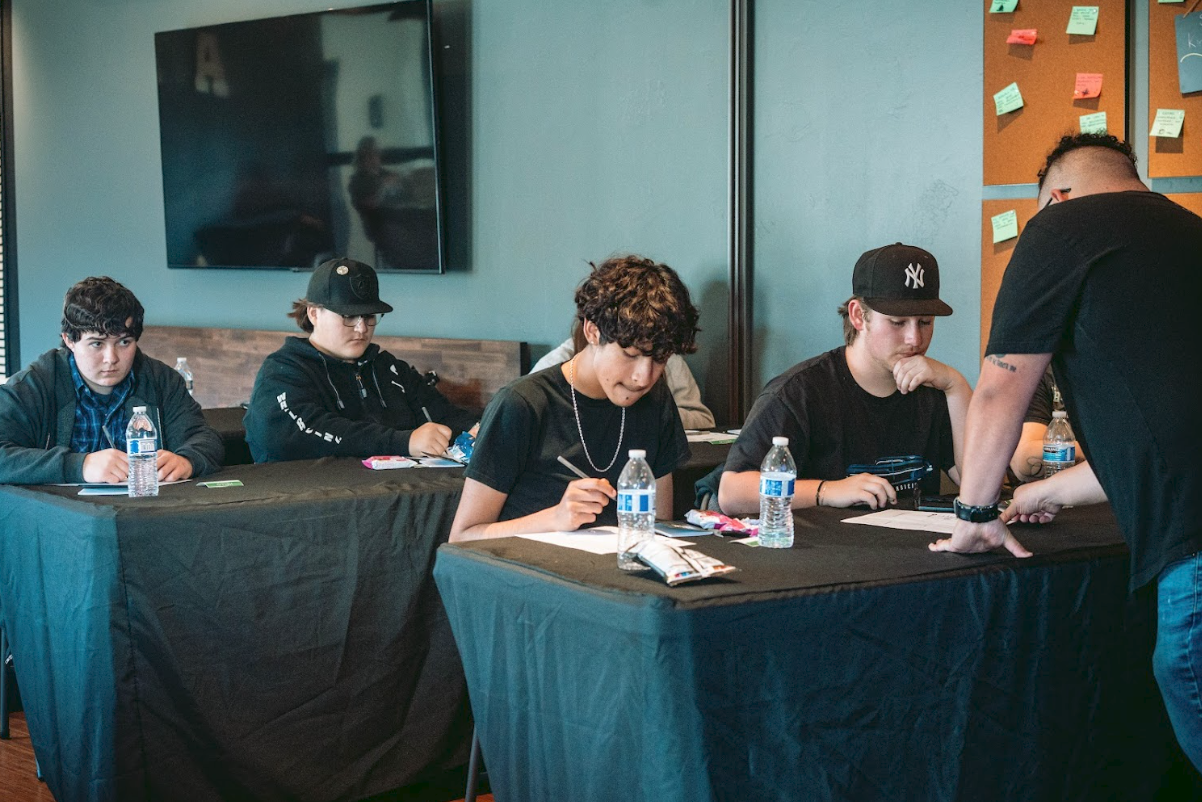 A group of young people sitting at a table in a classroom or workshop setting, with bottles of water and notebooks in front of them, listening to an instructor.
