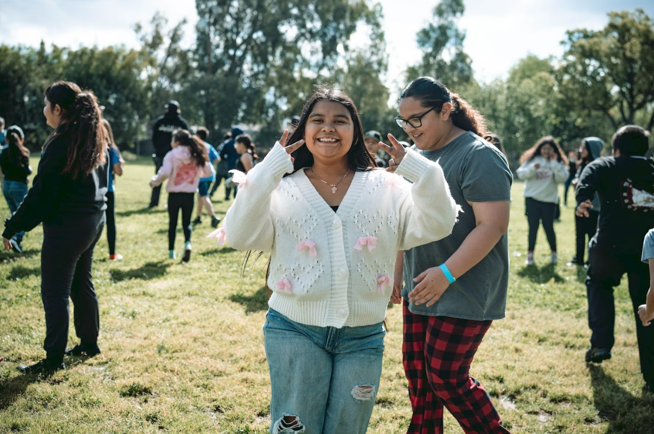 Smiling girl making peace signs with fingers, standing in a grassy park with other people in the background.