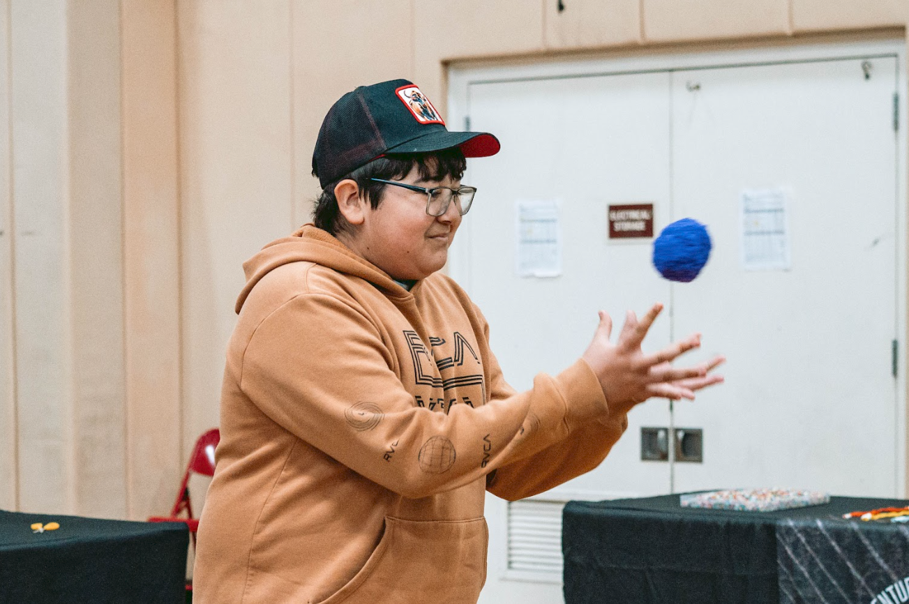 A young person wearing glasses, a black and red baseball cap, and an orange hoodie, is tossing a blue paper lantern in an indoor setting.