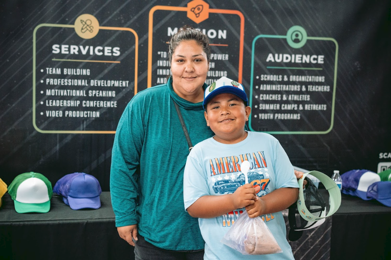 A woman and a young boy standing together at an event. The woman has dark hair tied back, is wearing a teal long-sleeve top, and has a lanyard around her neck. The boy is smiling, wearing a white T-shirt with a car graphic, a baseball cap, and holdin