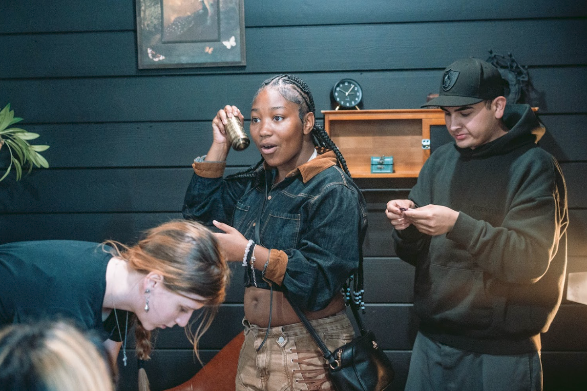 Three people, one woman and two men, are in a dark room with black wooden walls. The woman with light brown hair is leaning over, appearing to write or read something. The woman with braids and a denim jacket is speaking on a vintage microphone. The 