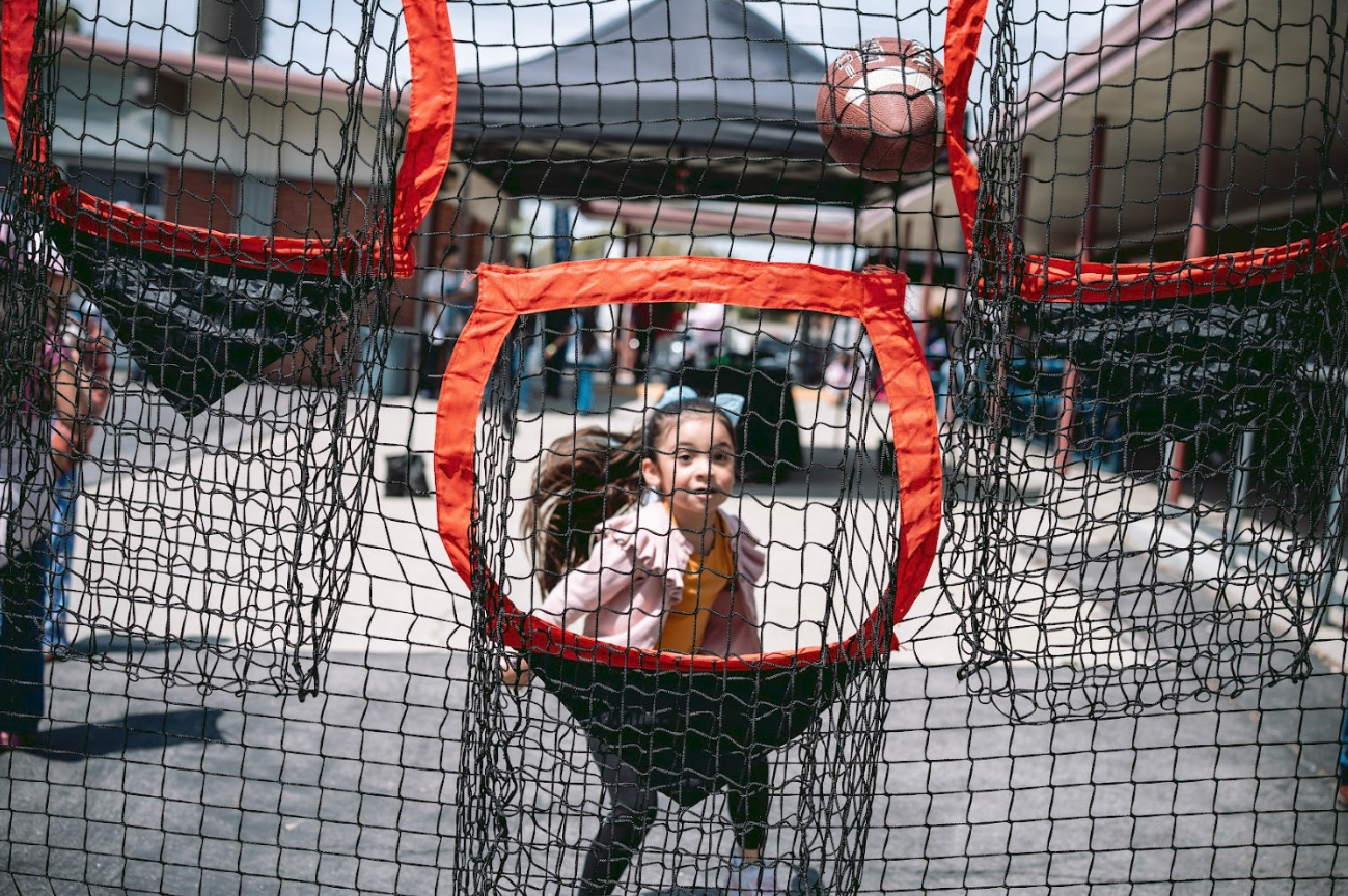 A young girl in a pink jacket and yellow top runs through a football toss game at an outdoor event. There is a net structure with bright orange trim and a football in the air inside the net. Other children and booths are visible in the background.