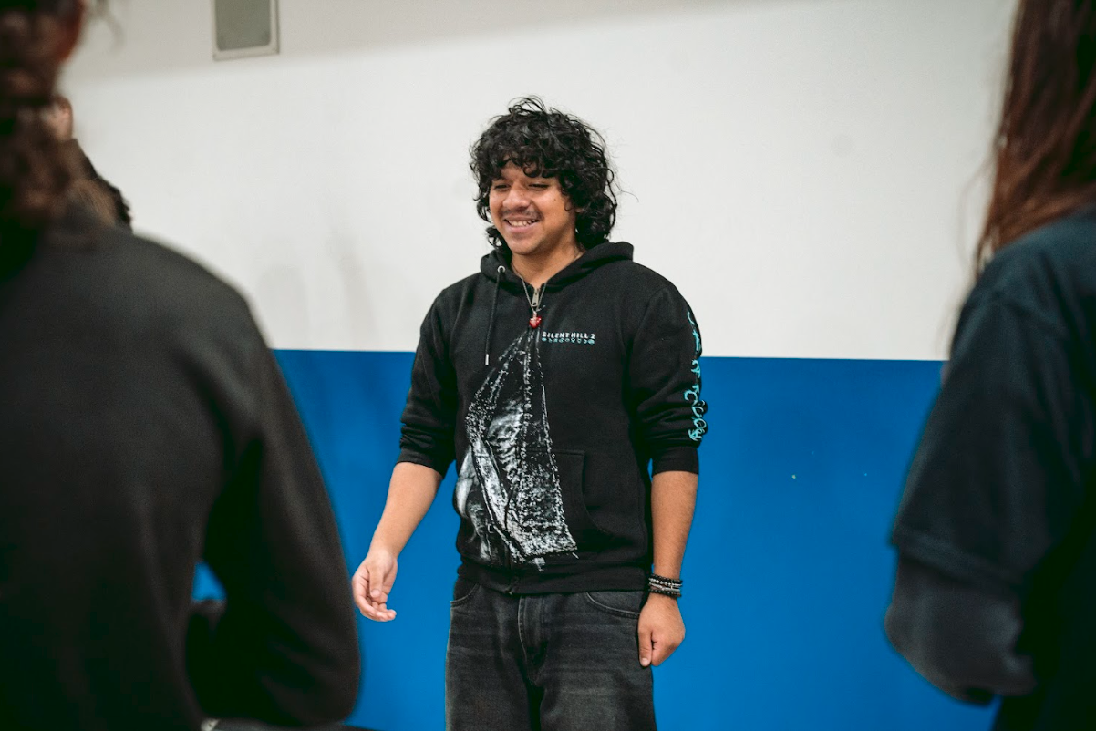 A young man with curly hair smiling and standing with others in an indoor setting.