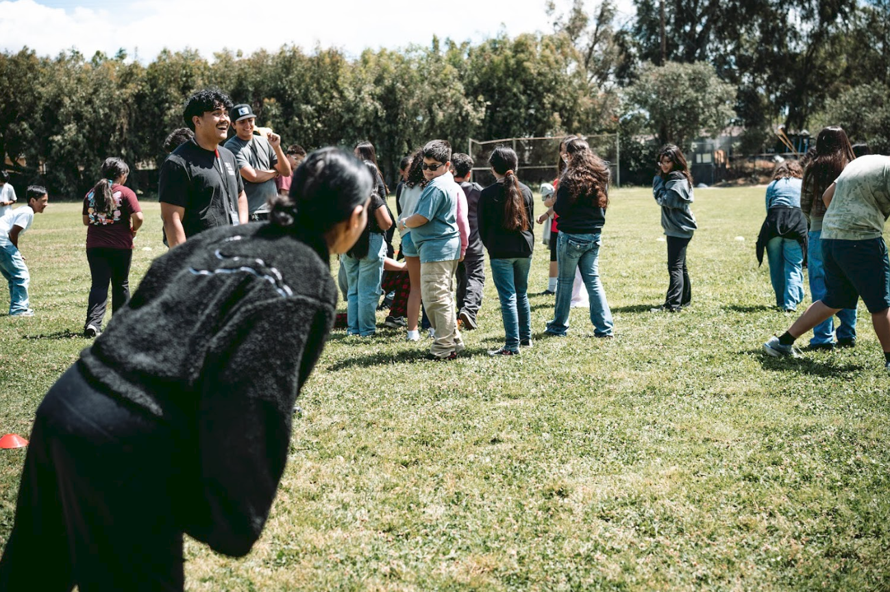 Group of children and teenagers on a grassy field participating in an outdoor activity, with some standing and others walking around, under a sunny sky with trees in the background.