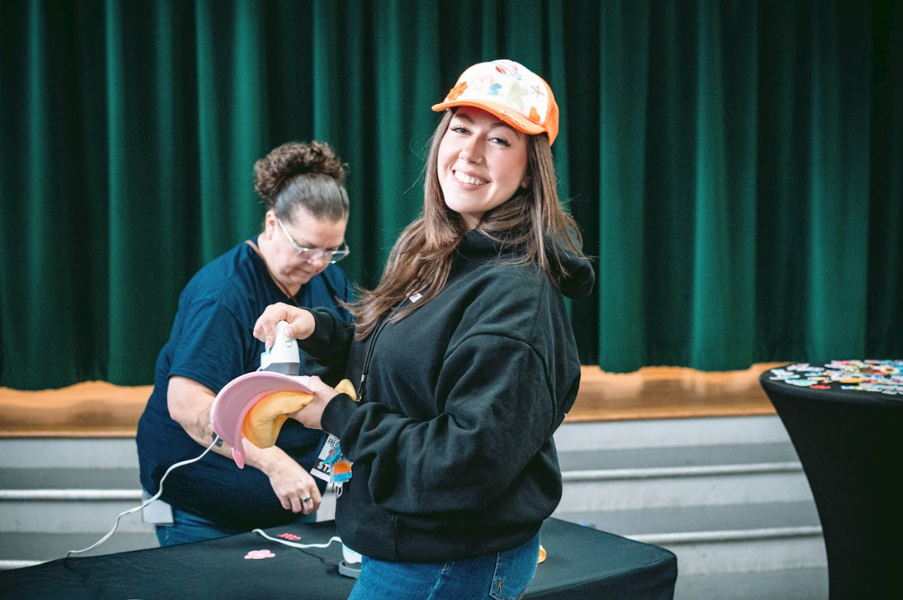 Young woman smiling and ironing a pink hat, standing in front of a green curtain, with another woman in the background.