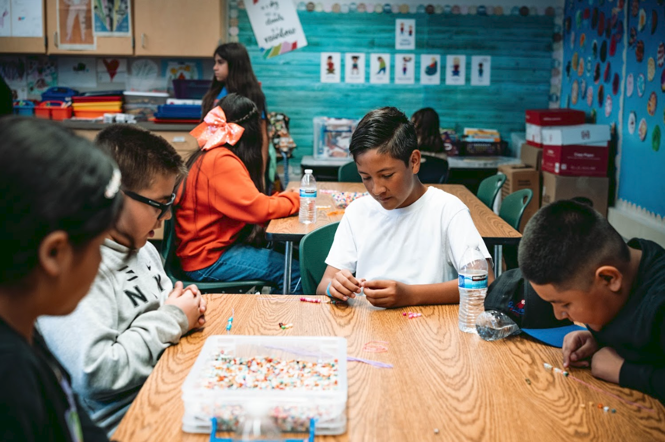 Children sitting at a classroom table decorating beaded jewelry, with colorful art and educational materials on the walls.