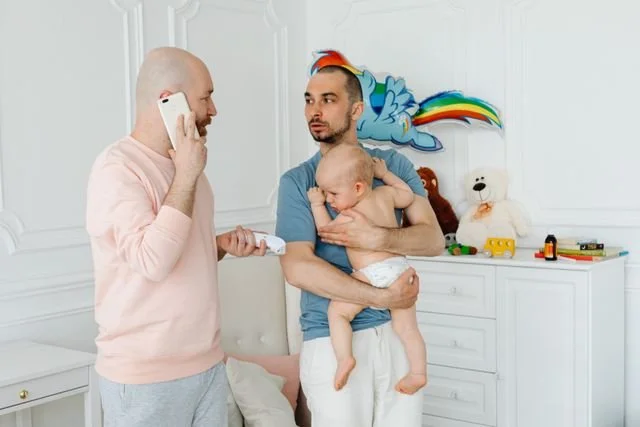 Two dads talk in a nursery while one holds a baby
