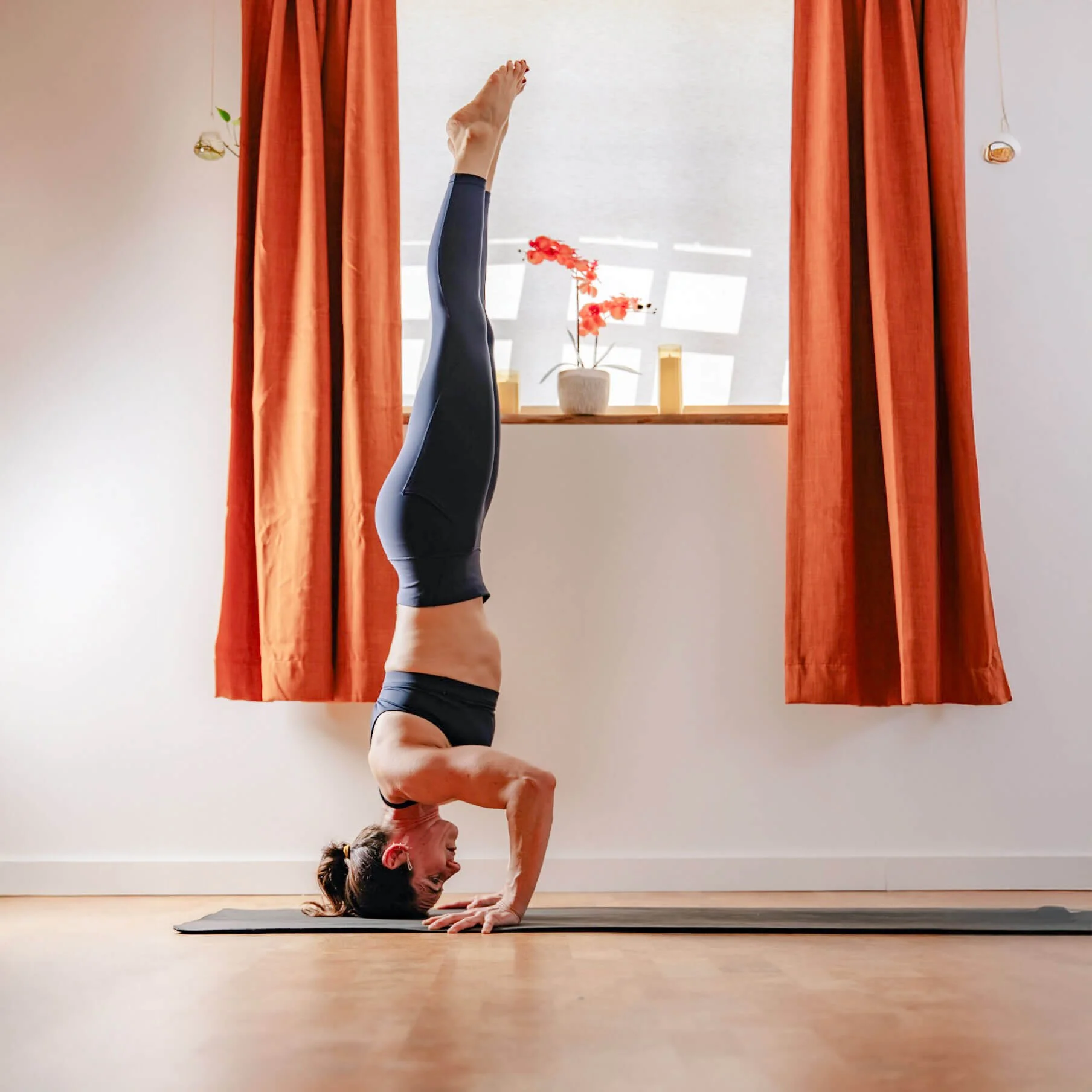 Two women practicing yoga together on a black yoga mat indoors; one woman is in a plank position, and the other is assisting her by holding her shoulder and smiling.