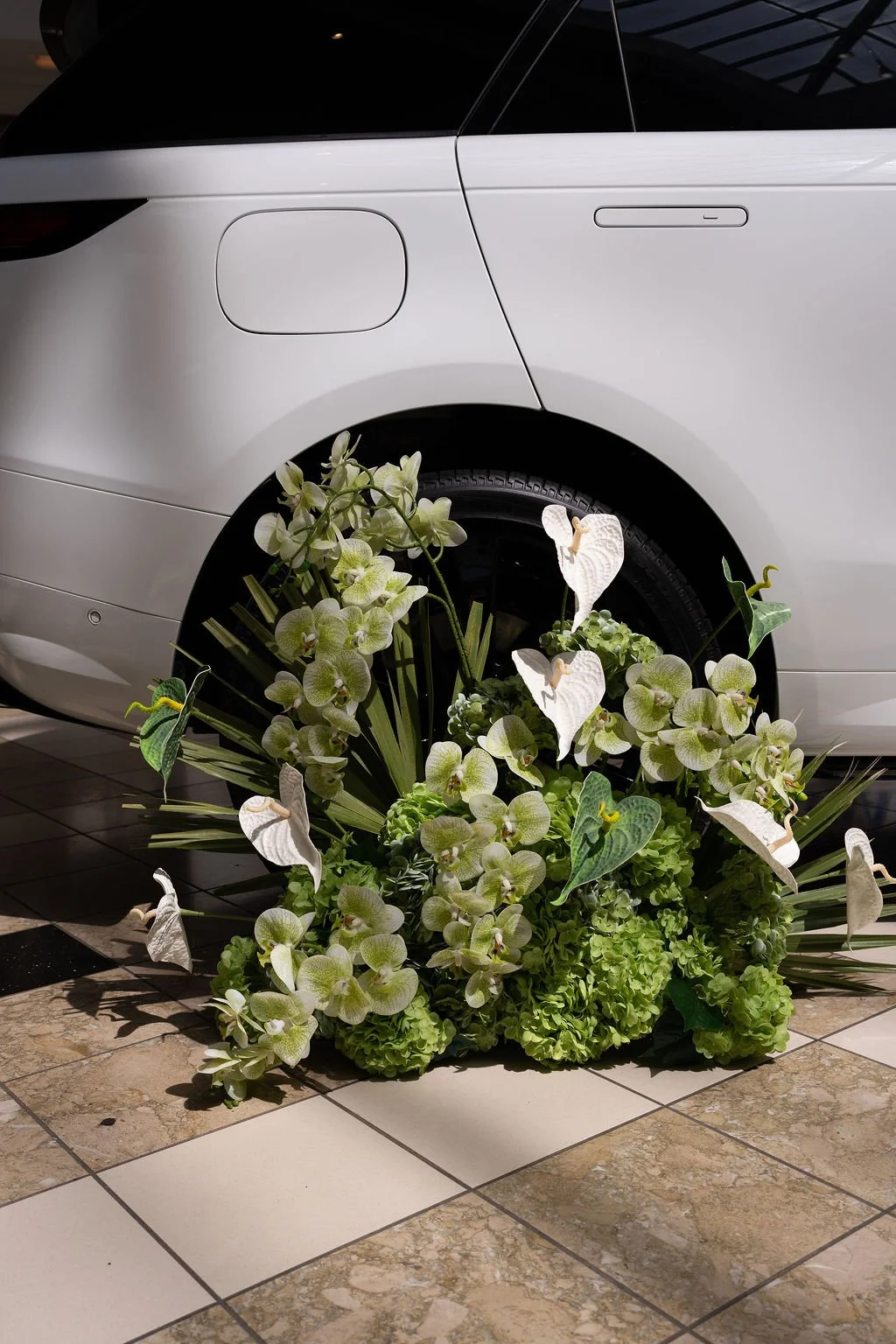 Editorial image of a luxury vehicle surrounded by a sculptural floral installation featuring green hydrangeas, orchids, and tropical foliage at an indoor brand activation