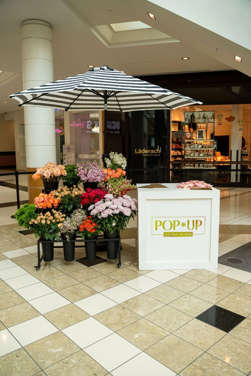 Editorial image of a floral pop-up installation at Pop-Up at the Plaza, featuring a striped umbrella, curated flower arrangements, and branded display counter inside a shopping center