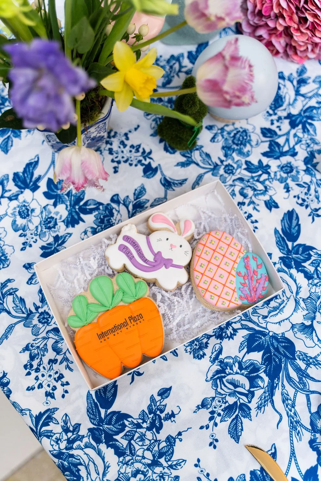 Editorial image of custom decorated cookies featuring International Plaza branding, styled in a gift box on a blue floral tablecloth for a seasonal brand activation