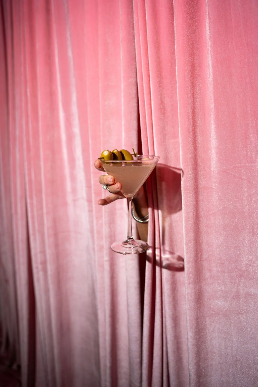 Editorial detail image of a hand holding a martini garnished with olives emerging through blush pink drapery, styled for a Palm Springs–themed gala event