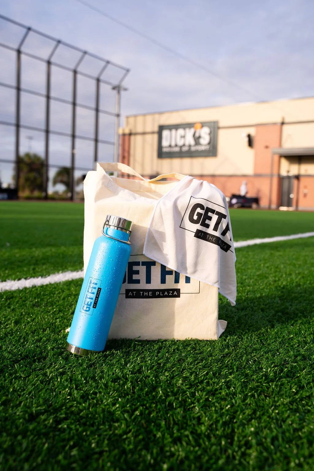 Editorial lifestyle photo of a smiling event guest on an outdoor turf field, carrying a ‘Get Fit at the Plaza’ branded tote bag and reusable water bottle during a fitness brand activation