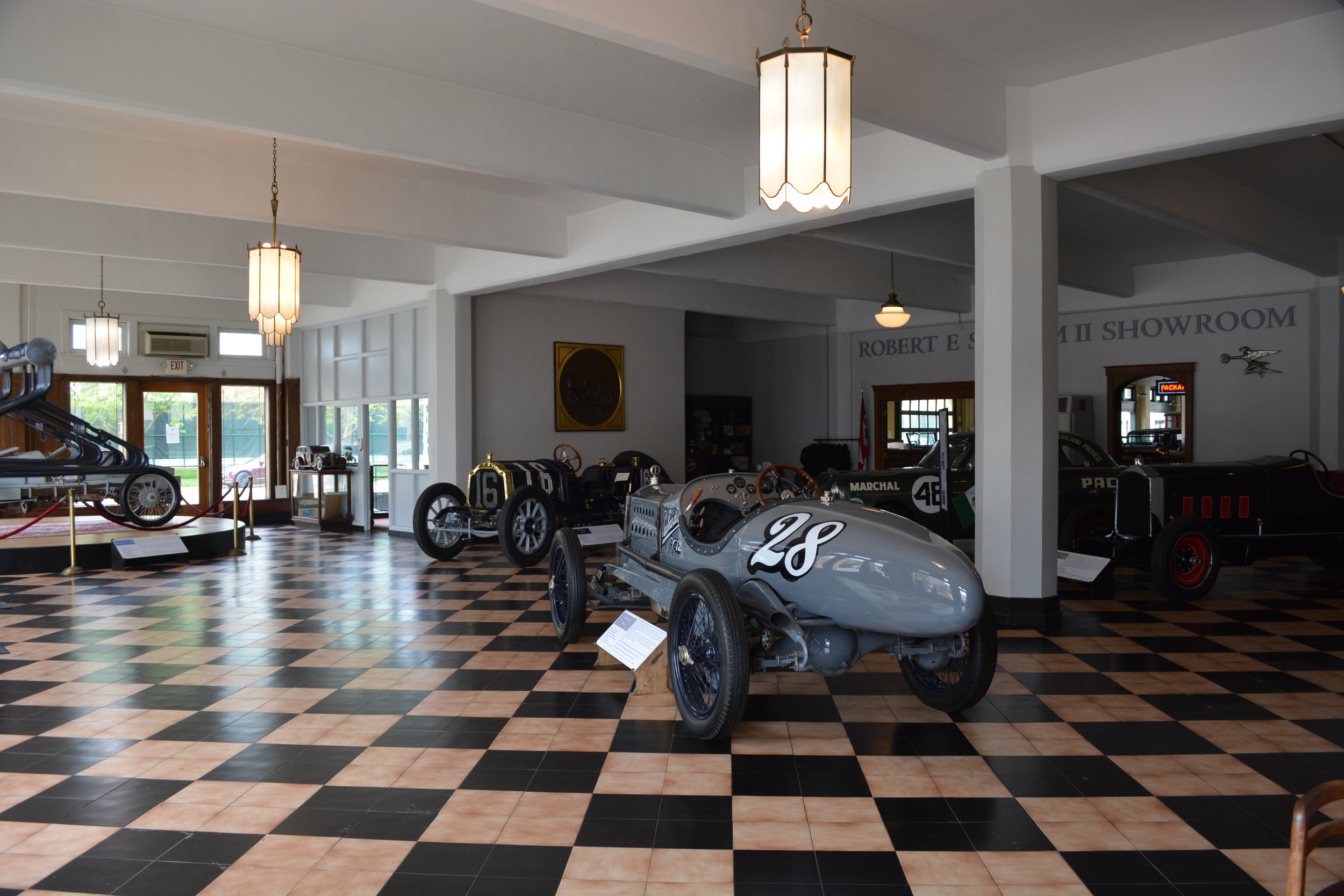 Interior of a vintage car museum displaying championship race cars, with checkered floor tiles, hanging light fixtures, and a glass entrance door.
