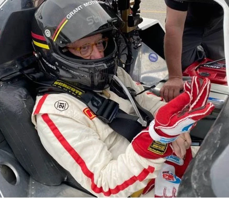 Race car driver in a helmet and racing suit sitting in a race car. The driver is holding racing gloves and appears to be preparing for a race.