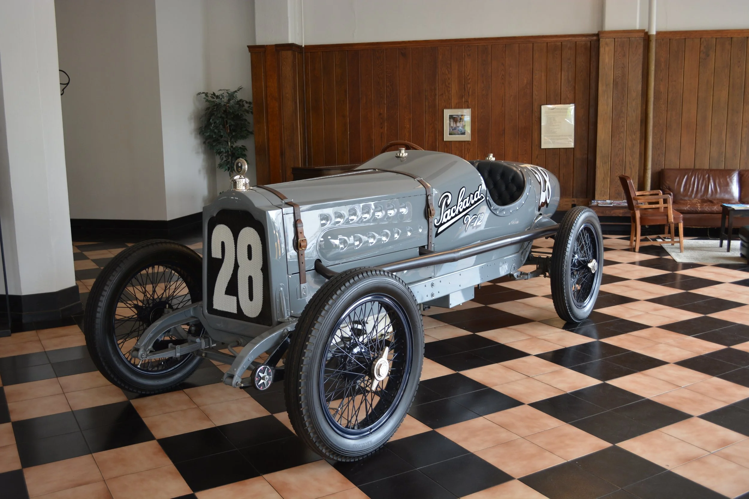 Vintage race car with the number 28, displayed indoors on a checkered black and tan tile floor, with wood-paneled walls and a leather couch in the background.