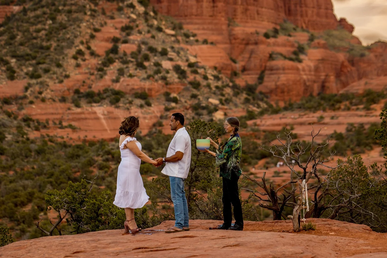 Sedona vow renewal couple on stunning red rocks with Bell Rock in the background. Officiant stands next to the couple, playing the crystal singing bowl. Couple holds hands, eyes closed.