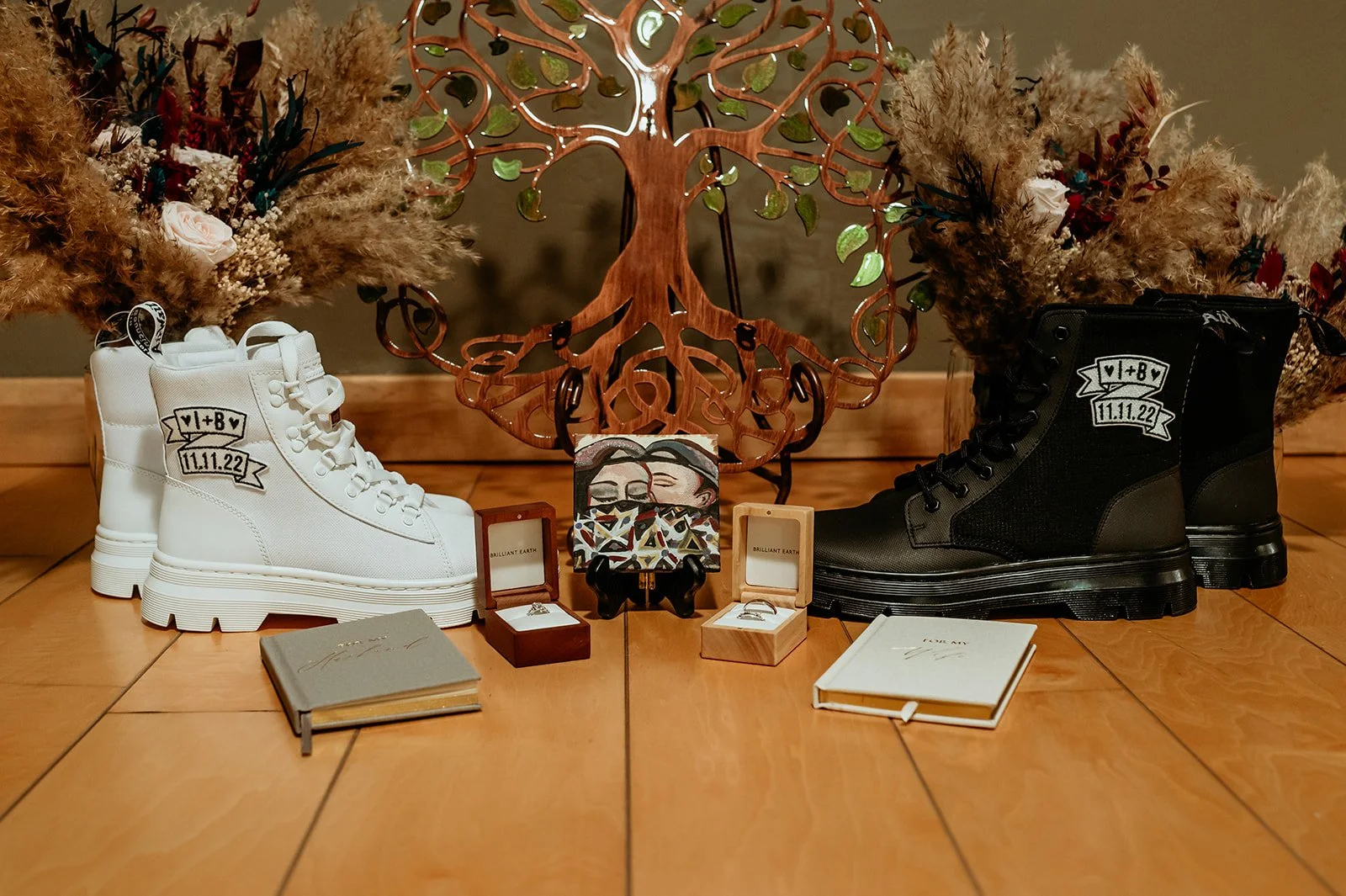 Bride and groom's hiking boots with dried flowers and metal tree art, wedding rings in boxes, and vow booklets. Bride's boots on left are white, groom's are black.