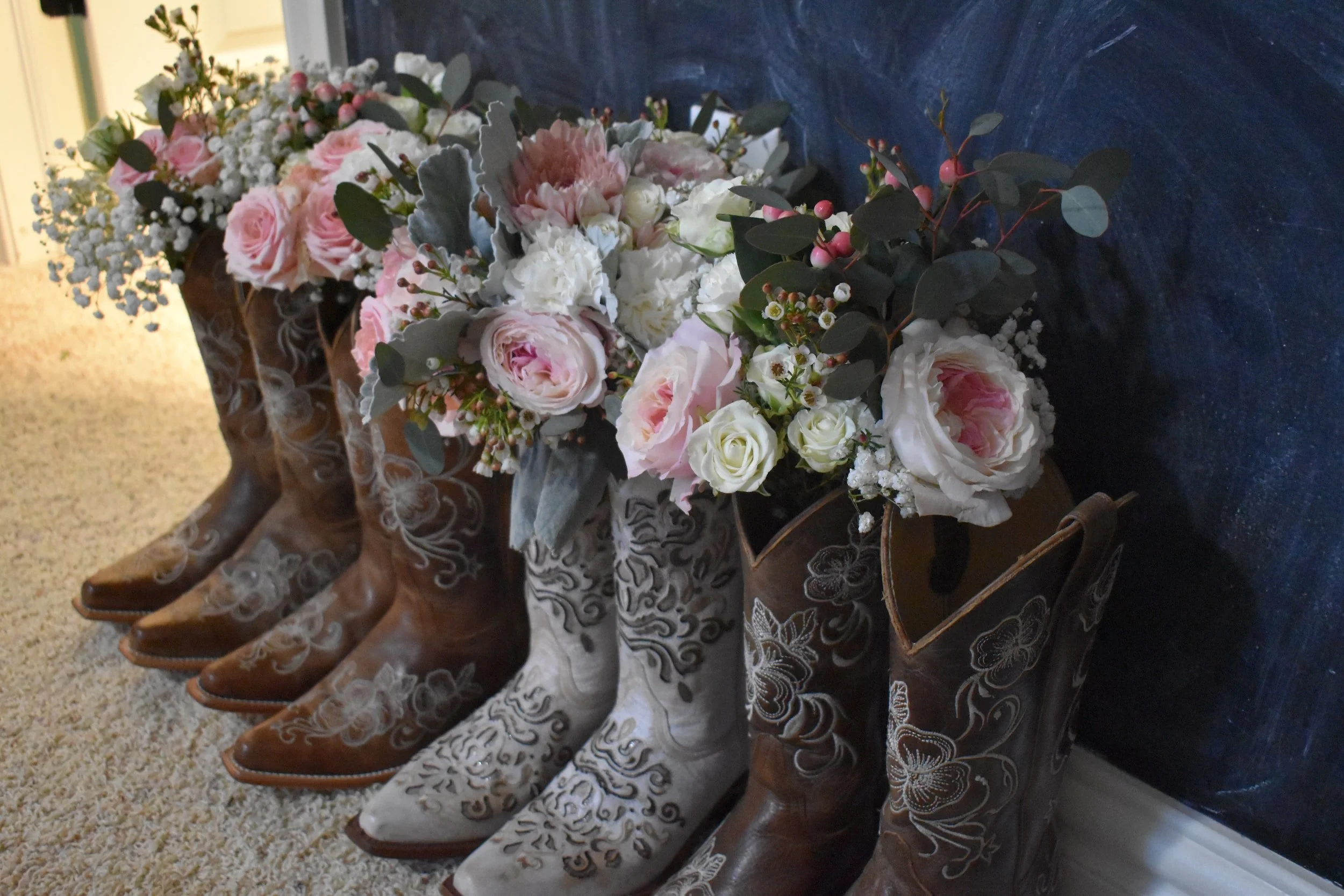 Row of four pairs of cowboy boots. Three pair are brown, and one is white and light grey. All have decorative flower stitching. Each boot has wedding flowers coming out the top..