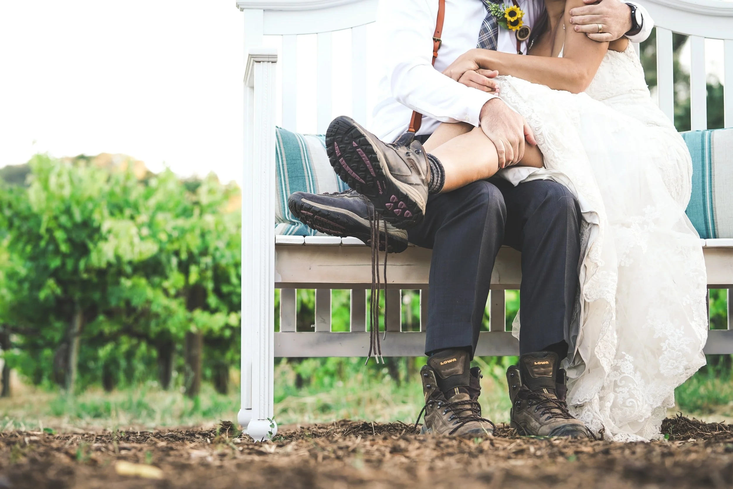 Legs of outdoor bride and groom. Bride sits on grooms lap. She wears hiking boots. The laces are untied and hand towards the ground. Groom also wearing untied hiking boots. Couple sits on light wood bench with trees in background.