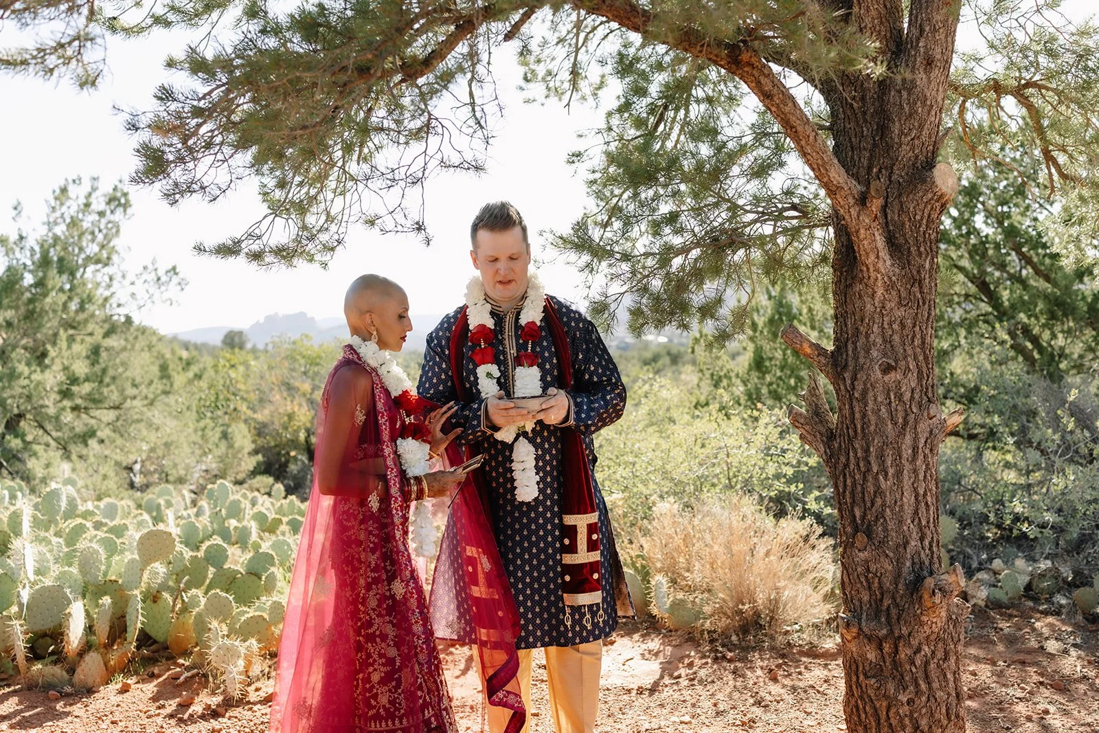 Outdoor wedding couple with dramatic background of Sedona's Cathedral Rock. Couple mostly faces one another with their backs mostly to the camera. Bride in elegantly simple white dress, groom in tailored suit.