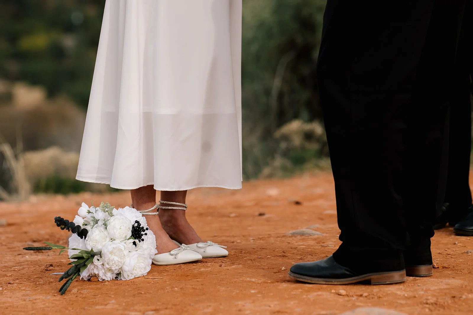 Legs of bride on left facing groom on right. White wedding bouquet at bride's feet. Bride's plain white dress hangs to her lower calf, and she wears white wedding shoes. Groom in black pants with black dress shoes.