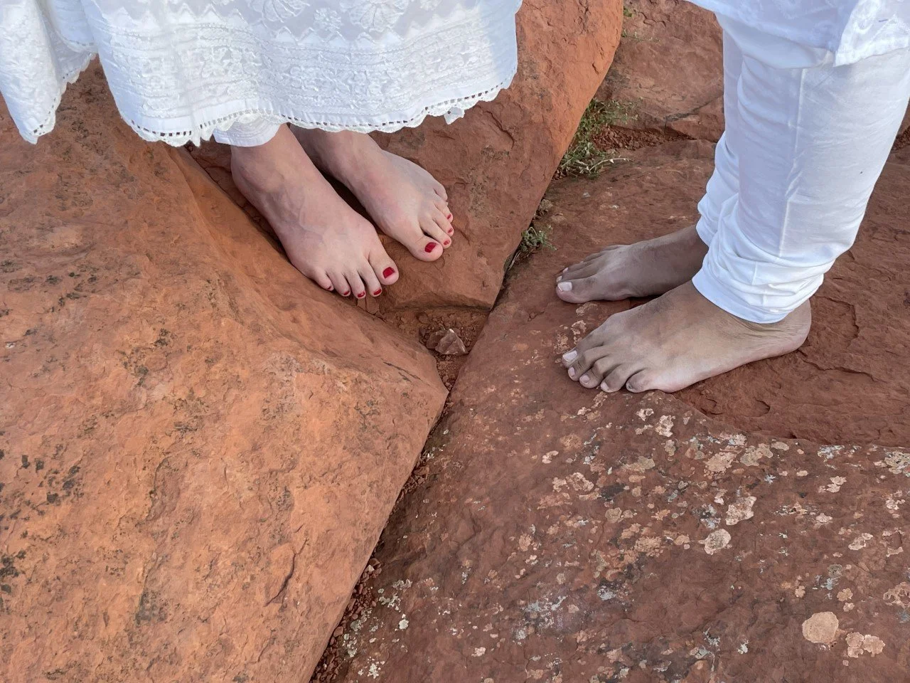 Couple's bare feet standing on Sedona red rocks. Bride on left has red painted nails. Hem of her white dress visible. Groom wears white pants. Their toes face one another.
