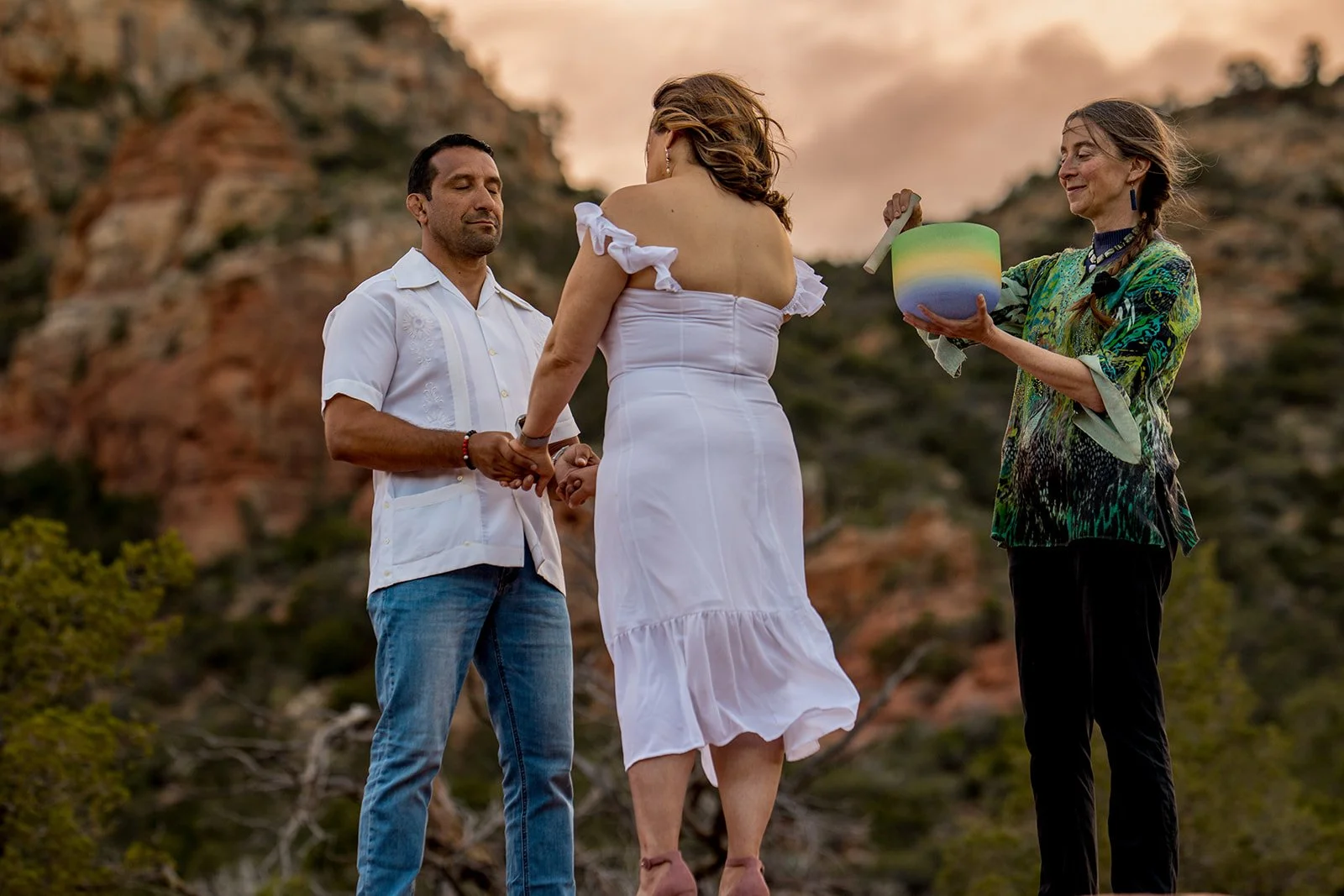 Sedona vow renewal couple, holding hands while officiant plays the crystal singing bowl. Husband's face is towards the camera, eyes closed. Red rocks with plants out of focus in the background.