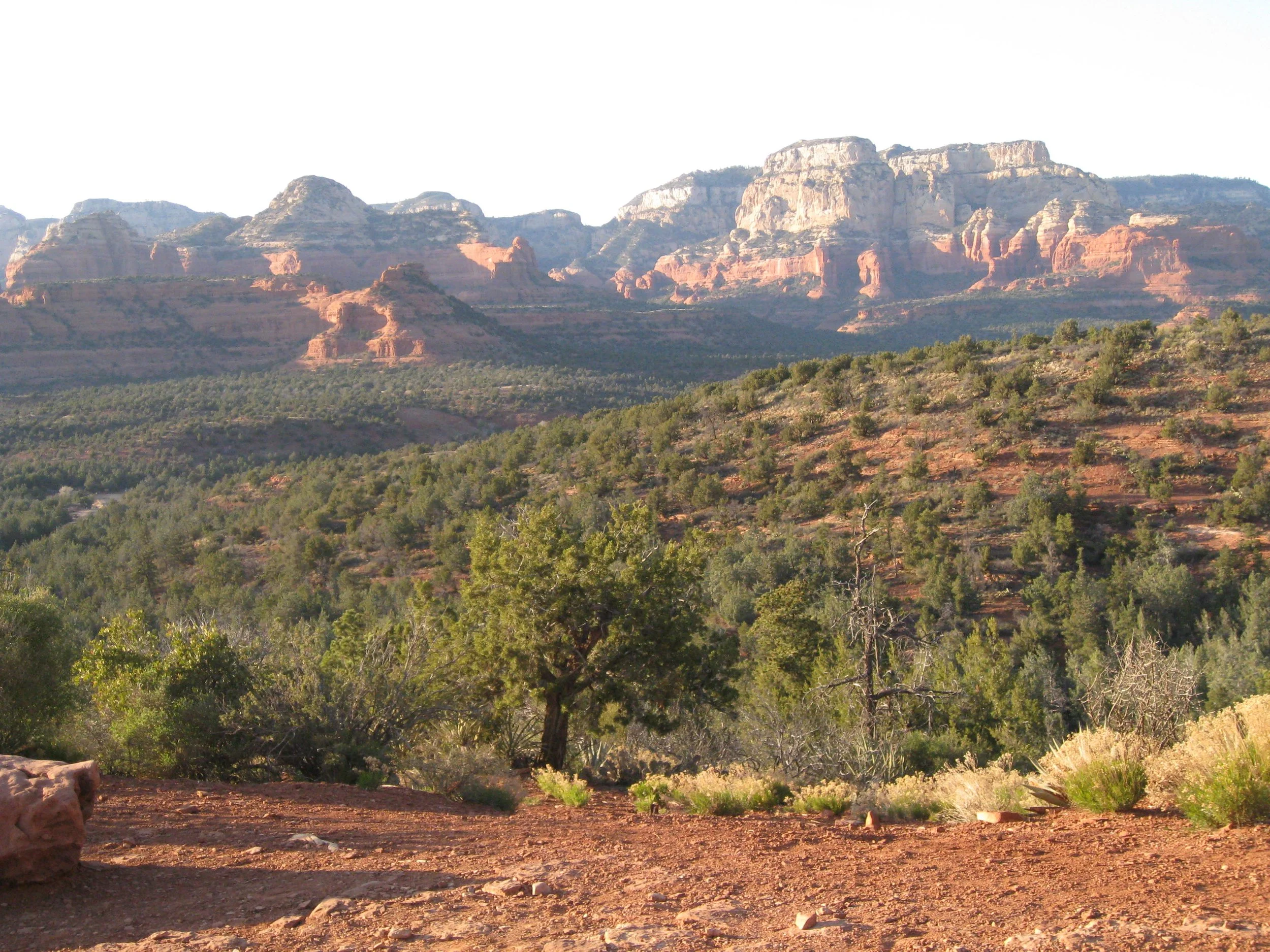 Landscape photo of Sedona. Flat red rock ground in foreground, beautiful red rock mountain in background. More flat area in between, with greenery.