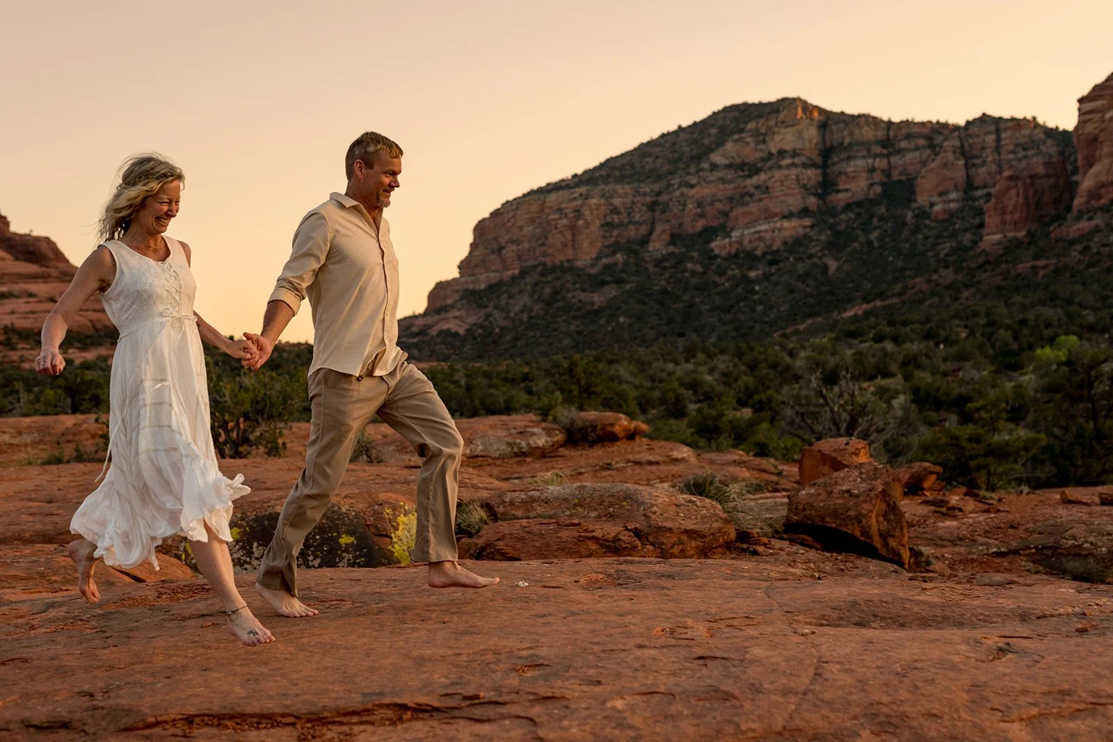 Barefoot Sedona elopement couple holding hands and running from left to right. Bride in casual white dress, groom in light casual brown pants and tan long-sleeved buttoned shirt. Sedona red rock landscape in background. Bride is laughing.