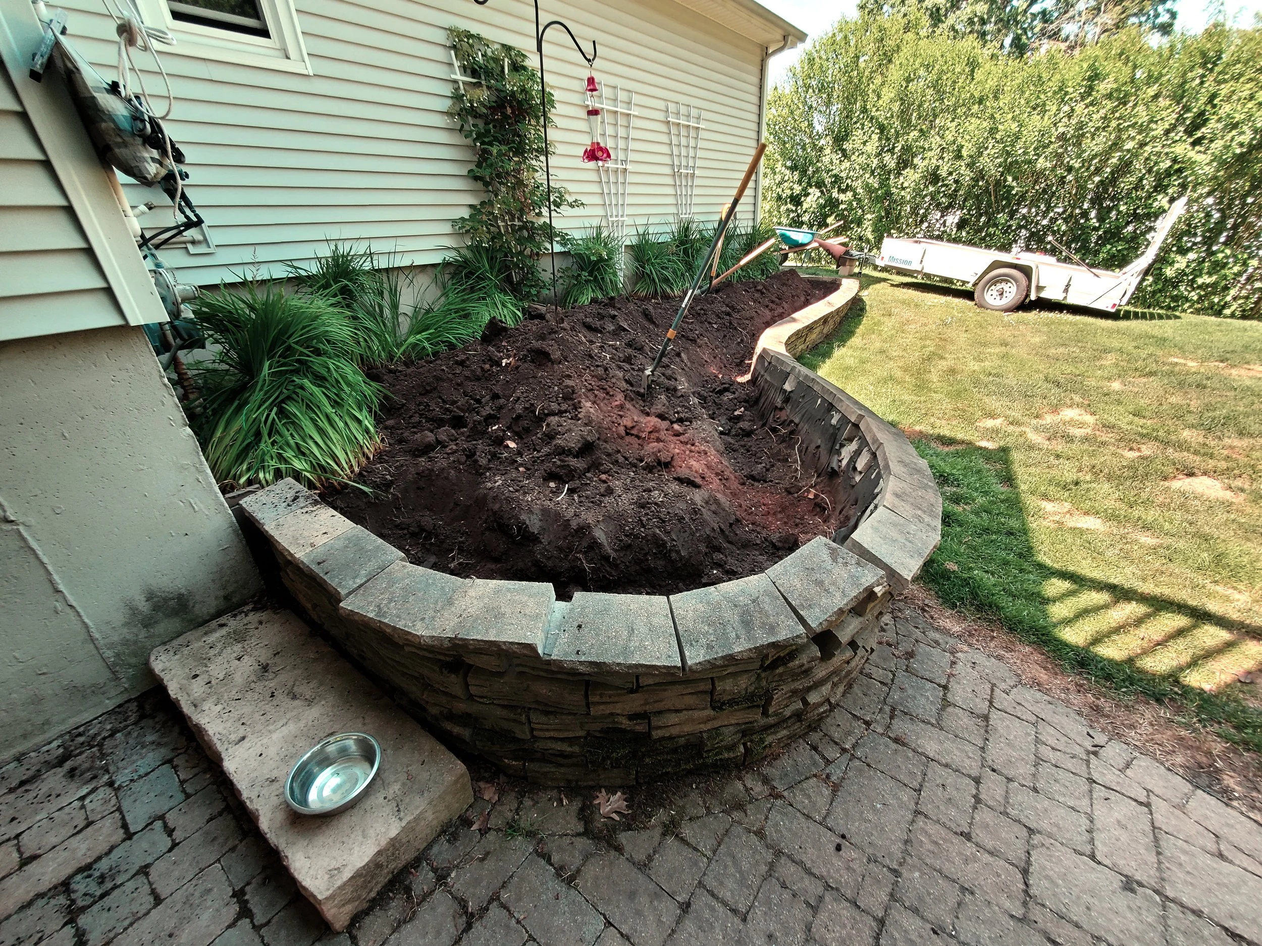 A partially built brick garden bed along a house wall, with gardening tools and plants nearby, and a lawn and trailer in the background.