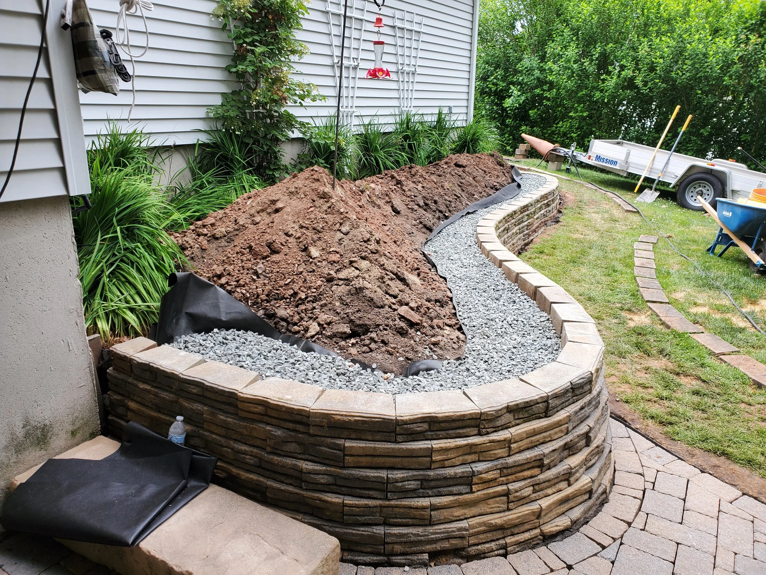 Ongoing backyard landscaping with a curved stone retaining wall, soil piles, gravel, and construction tools near a white house with green bushes.