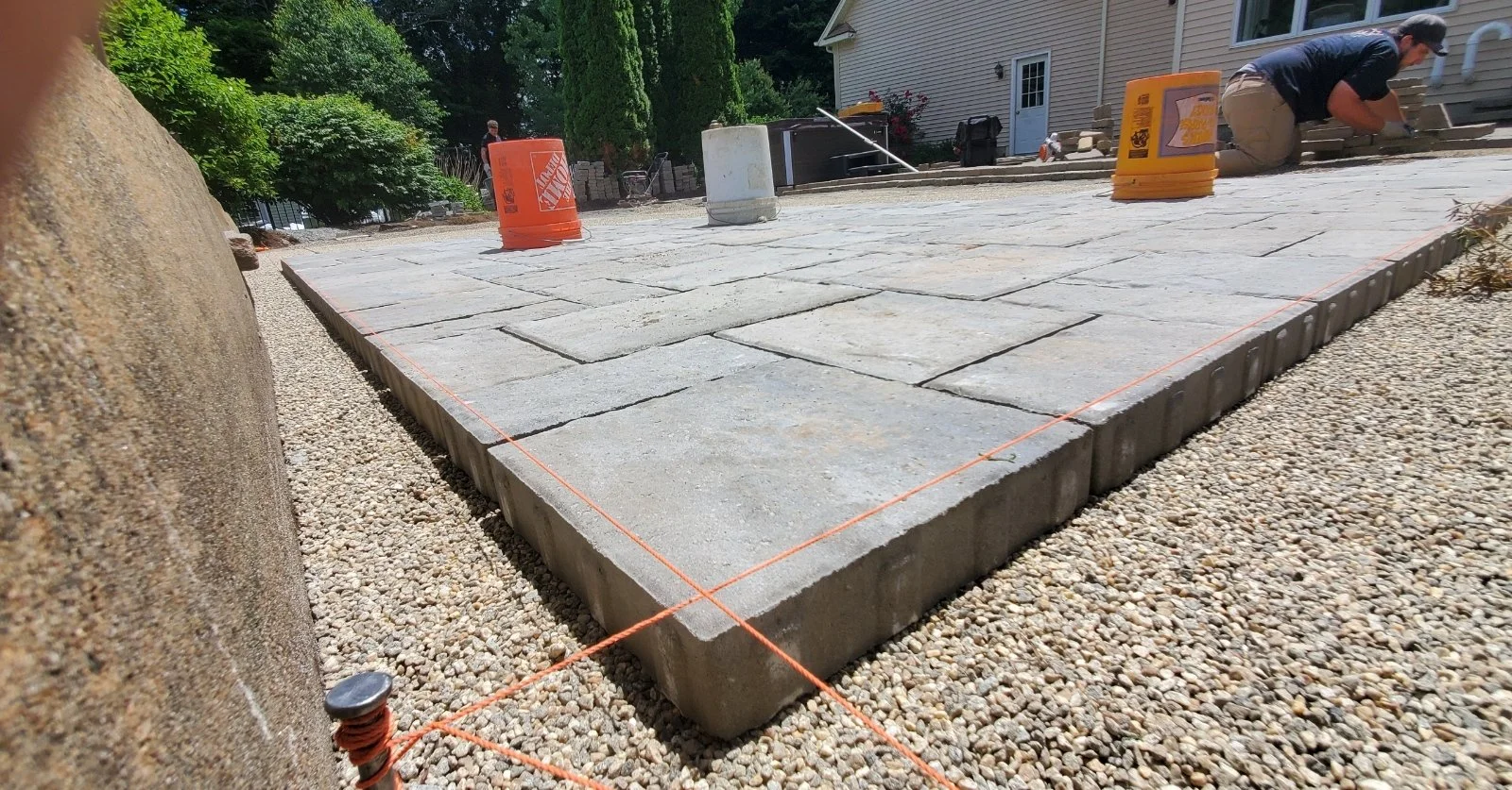 Person working on laying stone pavers on a patio with orange buckets and construction tools around.