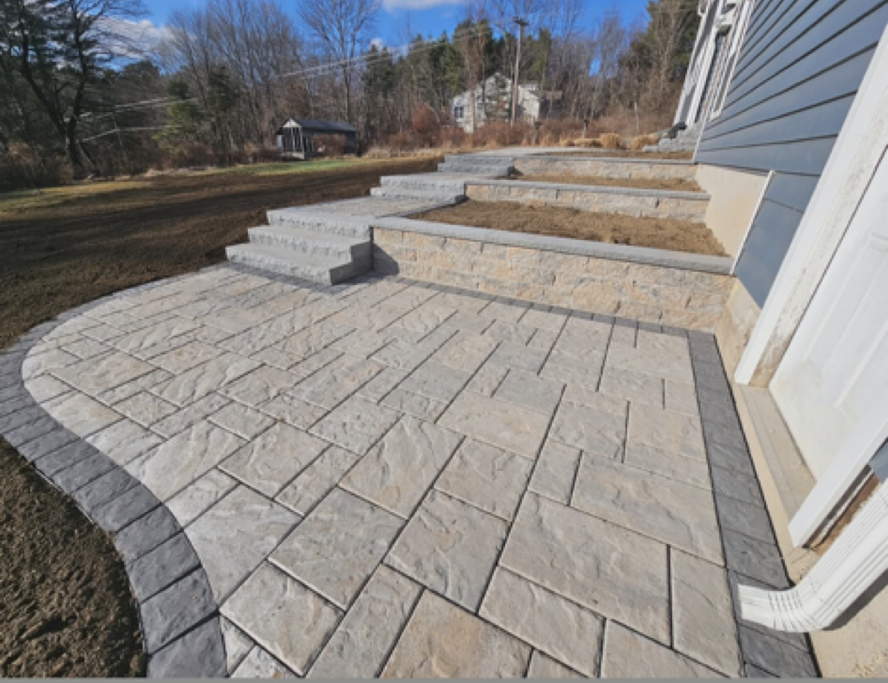 Newly installed stone patio and steps outside a house with a garden area and neighboring houses in the background.