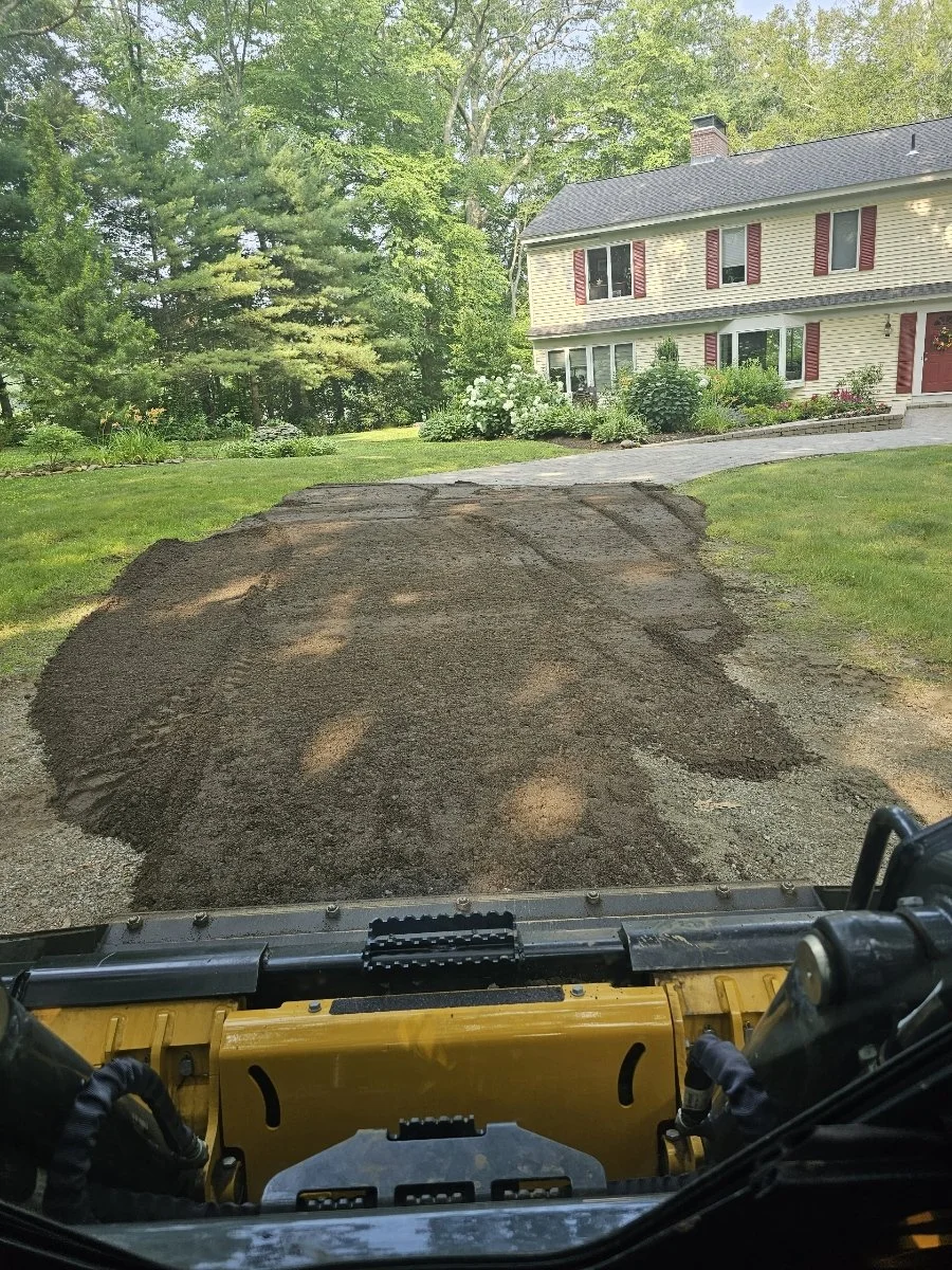 View from a construction vehicle looking at a freshly graded dirt driveway in front of a beige house with red shutters, surrounded by trees and green lawn.