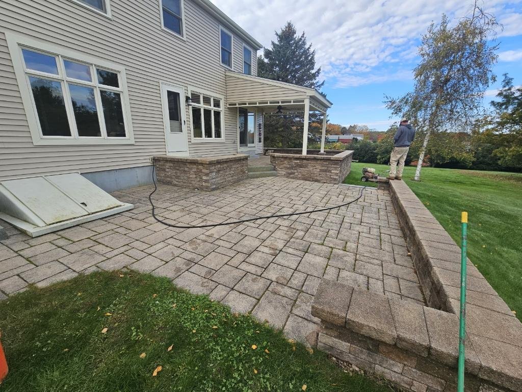 View of a partially completed brick patio extension outside a beige house, with a man working on the edge and a blue sky with some clouds.