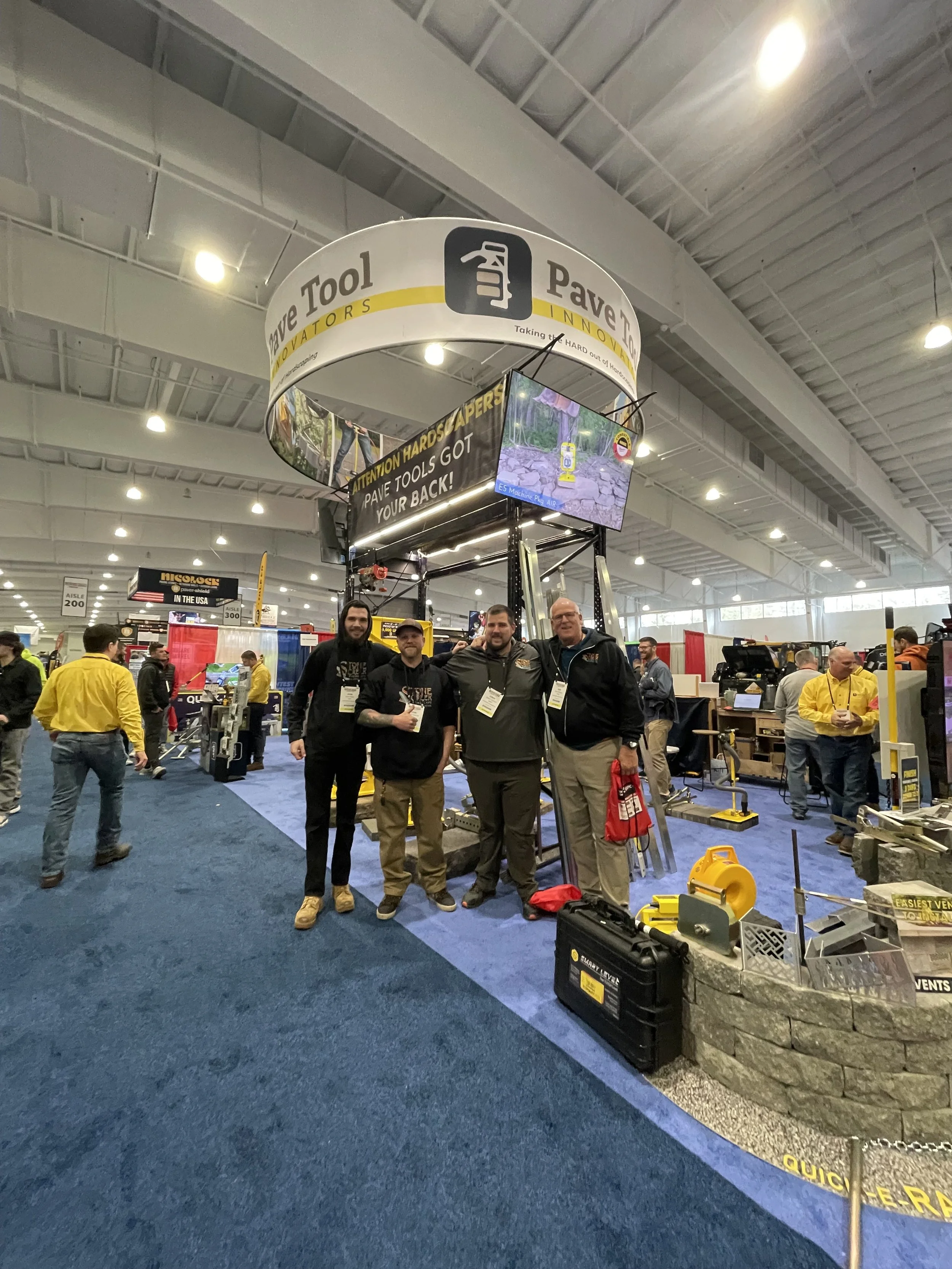 Four men stand together at a trade show booth for paving tools. The booth has a large circular sign overhead that reads "Pave Tool Innovators" and displays a small video monitor. Other vendors and attendees are visible in the background.