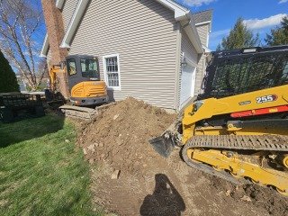 Construction equipment digging a trench next to a house with a pile of dirt nearby.
