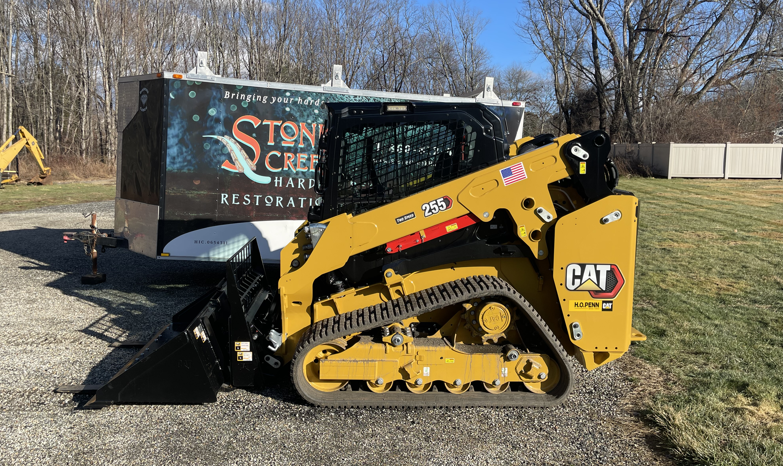 A yellow CAT compact track loader parked on a gravel surface with a black attachment in front. Behind it is a trailer with the text 'Stone Creek Hard Restoration,' and a background of trees with no leaves.