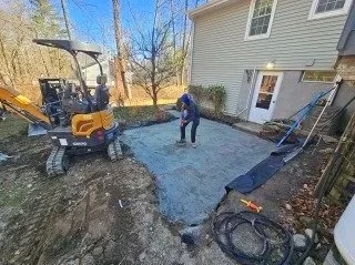 Person working on a freshly poured concrete patio in a backyard, with construction equipment and tools nearby.
