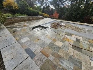 A damaged stone pathway with a large hole and exposed wiring, surrounded by trees and a stone wall.
