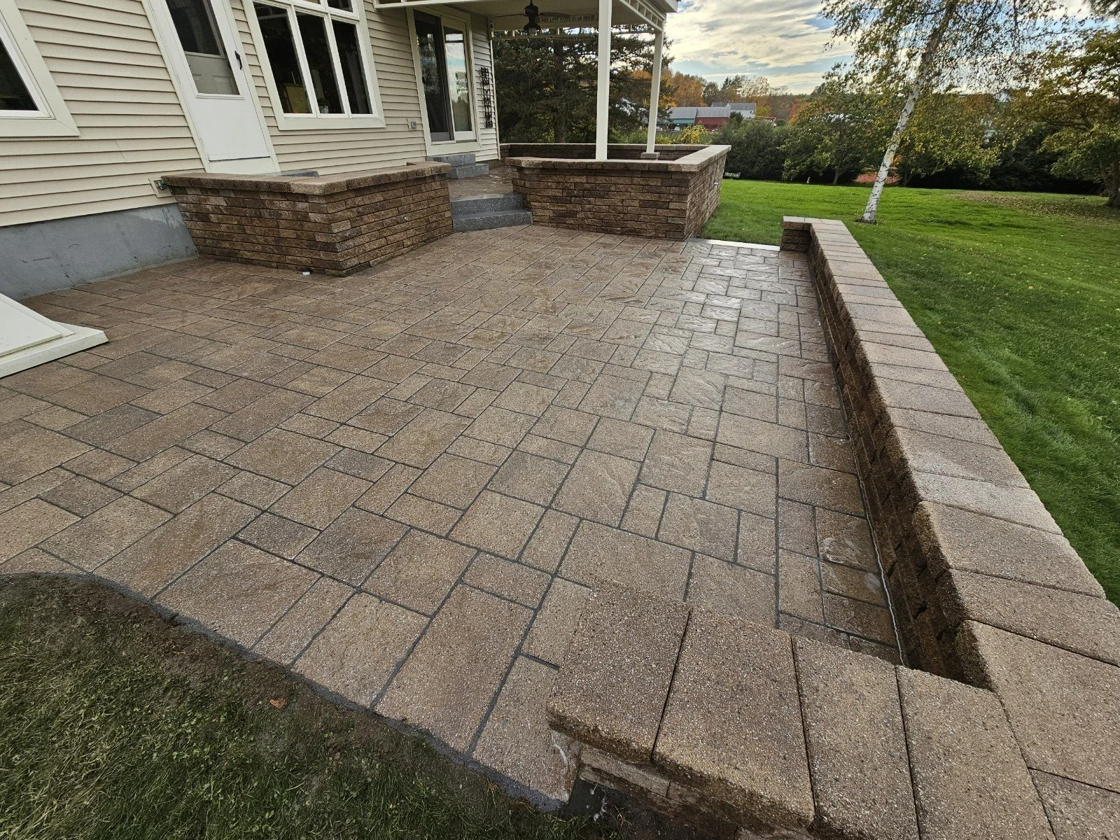 Photo of a newly paved backyard patio with brick retaining walls, stairs leading to the house door, and surrounding grassy yard with trees.