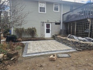 Backyard patio area with stone pavers, small tree, and a door leading to the house. Part of a deck is visible on the right.