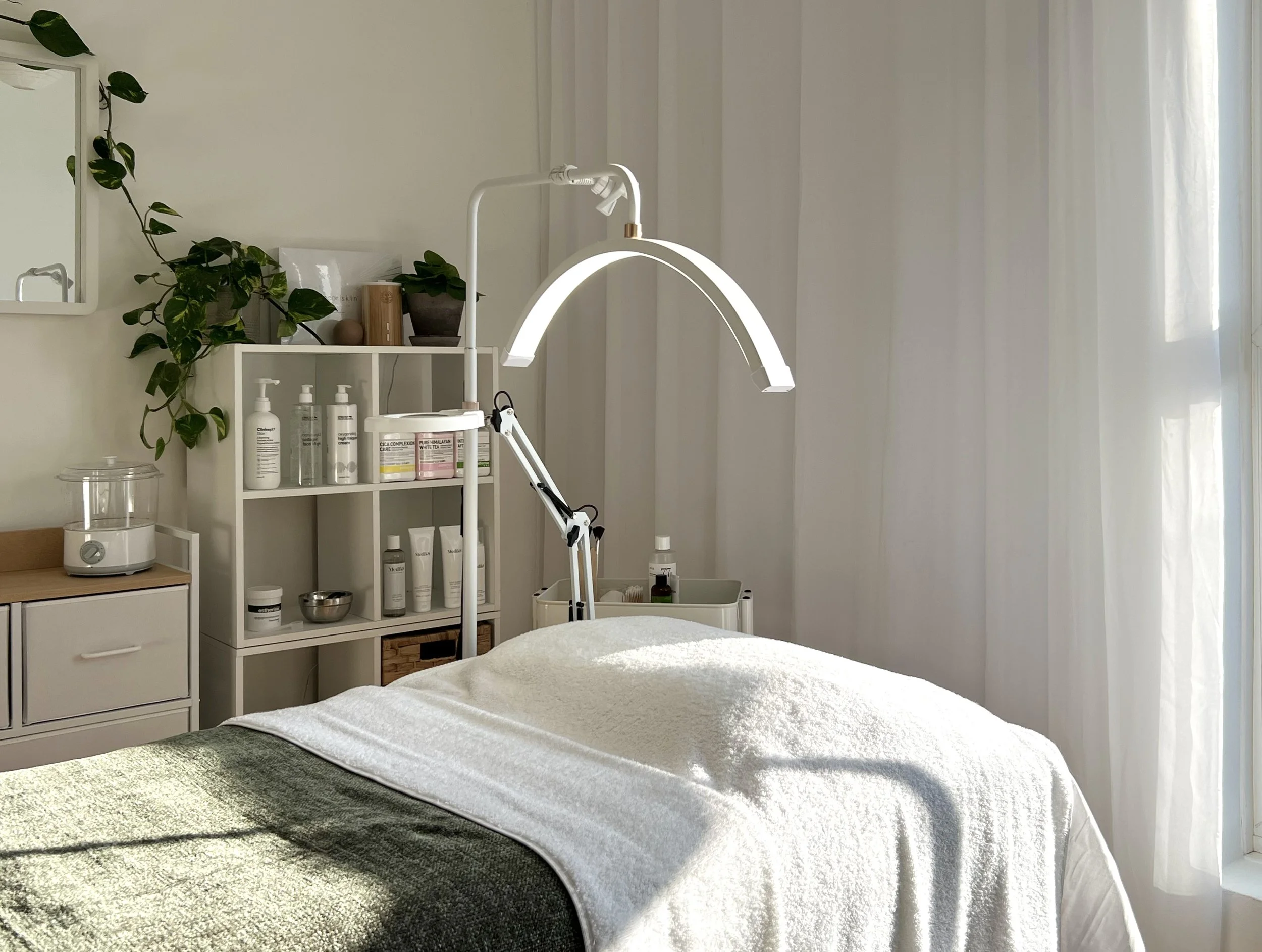 A clean, minimalist treatment room with a massage bed covered in white and dark green towels, a bright overhead lamp, and shelves with skincare products and plants.