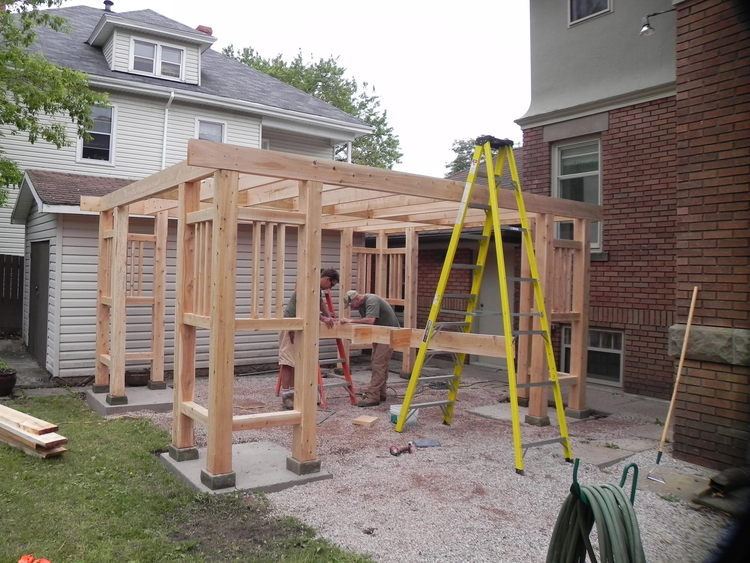Construction site of a wooden frame structure near a house, with two people working and a yellow ladder in the foreground.