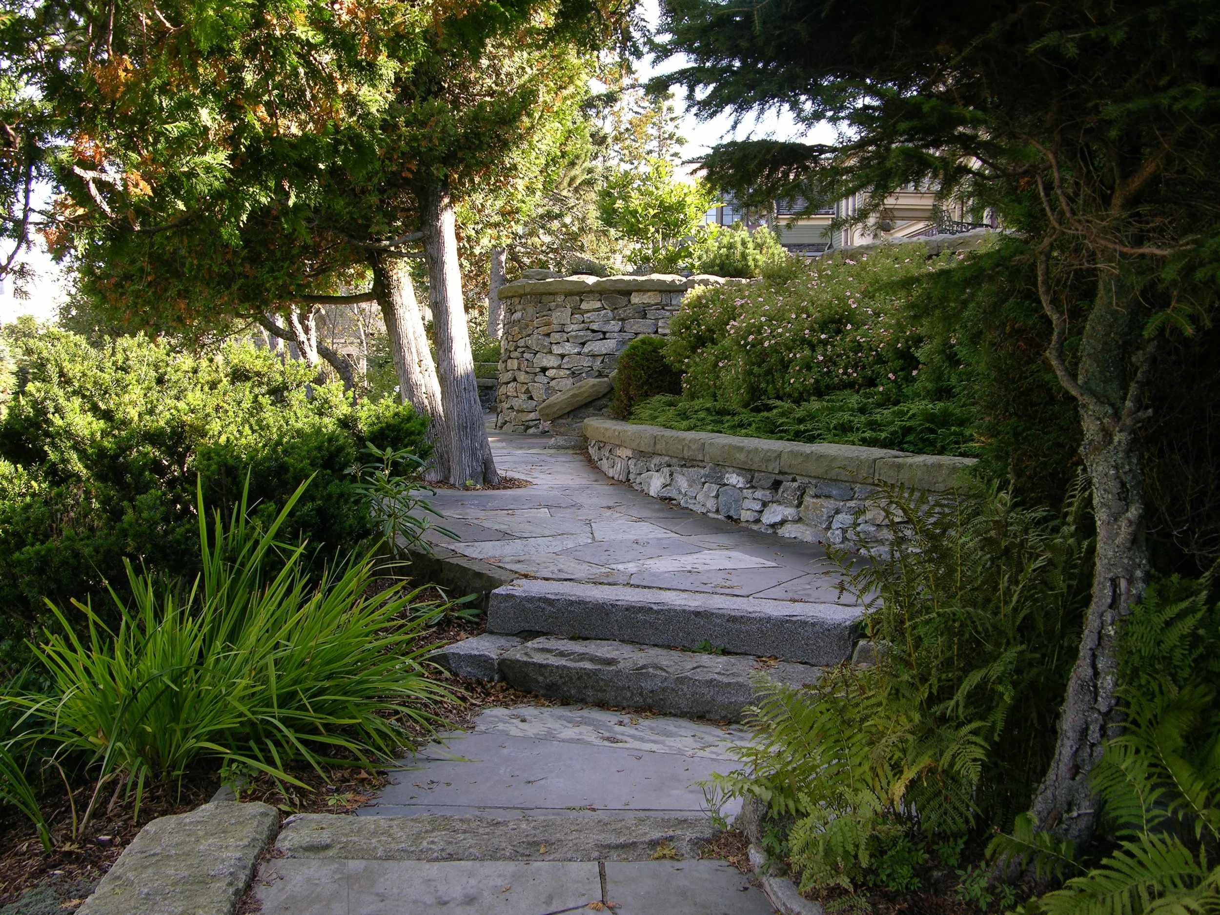 Stone pathway surrounded by lush greenery and trees, with stone walls and garden plants.