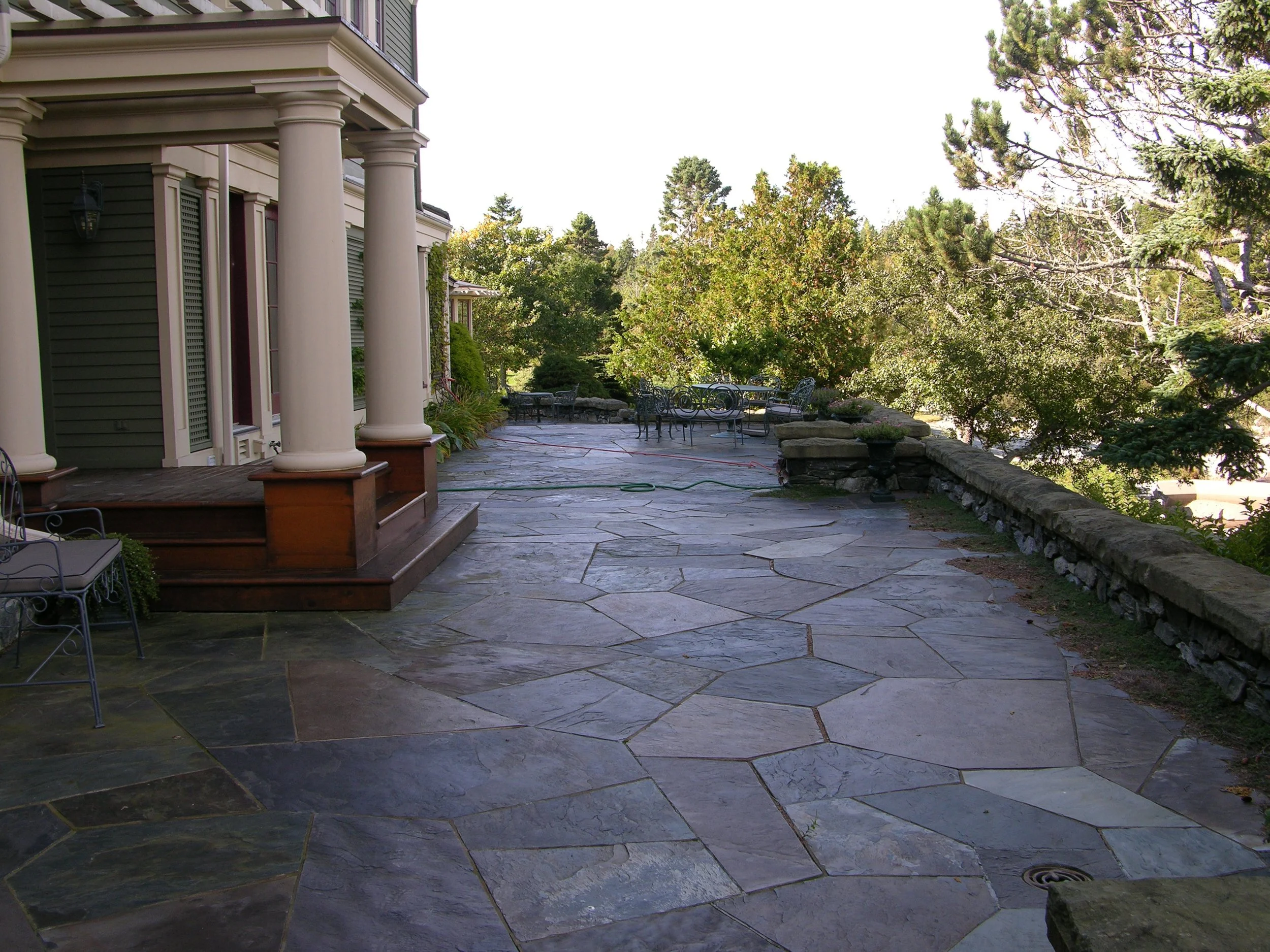 Stone patio with decorative seating, flanked by columns and trees, adjacent to a house.